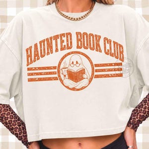 Puede incluir: Una camiseta corta de color blanco roto con un gr&aacute;fico naranja desgastado que dice "HAUNTED BOOK CLUB" en fuente arqueada. El dise&ntilde;o incluye un fantasma con gafas, leyendo un libro y sosteniendo una taza, flanqueado por rayas horizontales.