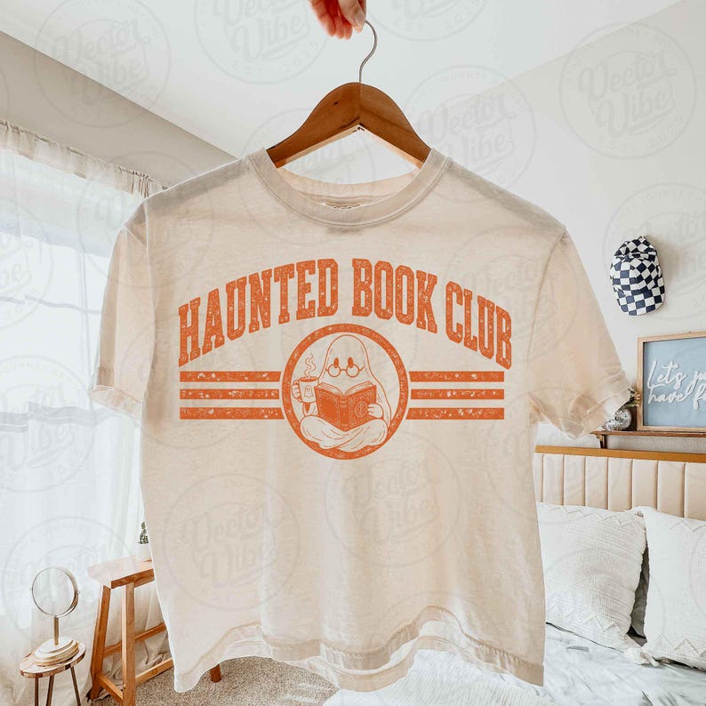 Puede incluir: Una camiseta de manga corta color crema con un gr&aacute;fico naranja que incluye el texto "HAUNTED BOOK CLUB" sobre una ilustraci&oacute;n circular de un fantasma con gafas, sosteniendo una taza humeante y leyendo un libro abierto.