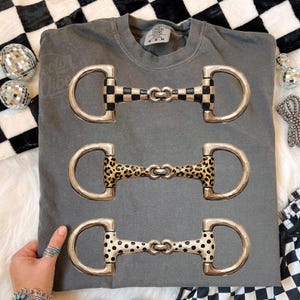 Puede incluir: Sudadera gris Comfort Colors con tres diseños decorativos de bocados de caballo. Las bocas tienen anillos metálicos plateados y centros estampados: cuadros blancos y negros, estampado de leopardo y lunares negros.
