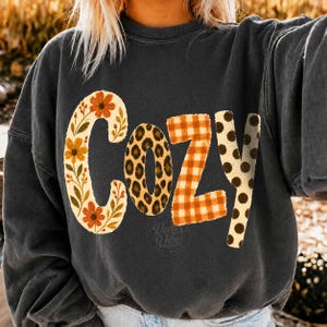 Puede incluir: Una sudadera de cuello redondo gris carb&oacute;n con la palabra COZY impresa en letras grandes. Cada letra tiene un patr&oacute;n distinto: la C es floral con flores naranjas, la O es de leopardo, la Z es de cuadros naranjas y la Y tiene lunares marrones.