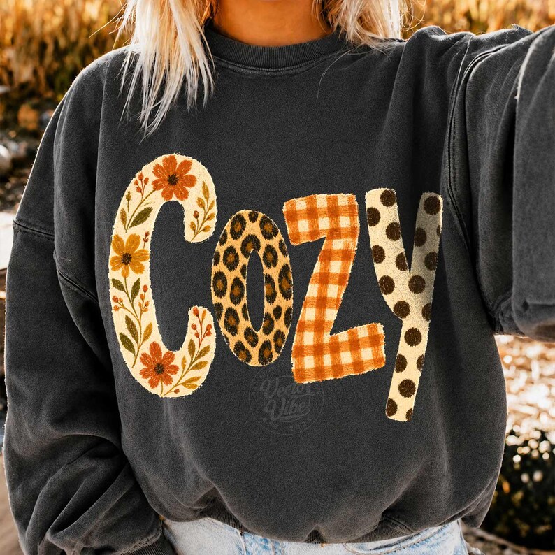 Puede incluir: Una sudadera de cuello redondo gris carb&oacute;n con la palabra COZY impresa en letras grandes. Cada letra tiene un patr&oacute;n distinto: la C es floral con flores naranjas, la O es de leopardo, la Z es de cuadros naranjas y la Y tiene lunares marrones.