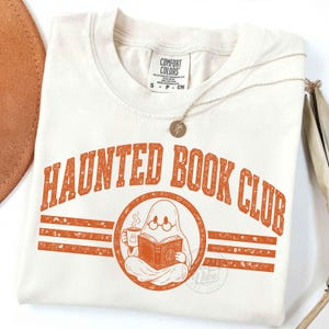 Puede incluir: Una camiseta de color crema con el texto naranja "HAUNTED BOOK CLUB" en una fuente universitaria. Debajo del texto hay un gr&aacute;fico circular de un fantasma con gafas redondas, sosteniendo una taza humeante y leyendo un libro abierto.