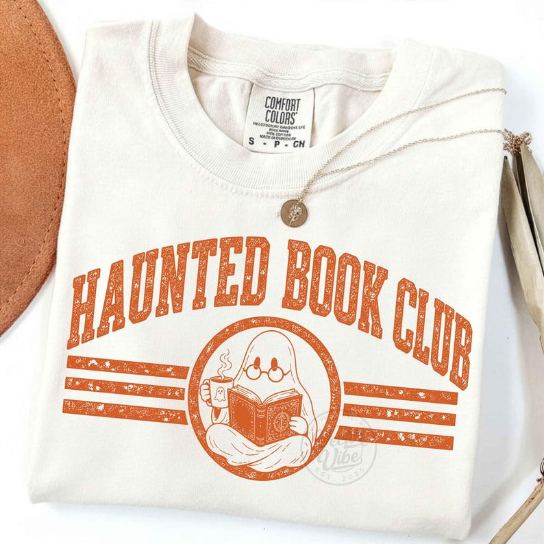 Puede incluir: Una camiseta de color crema con el texto naranja "HAUNTED BOOK CLUB" en una fuente universitaria. Debajo del texto hay un gr&aacute;fico circular de un fantasma con gafas redondas, sosteniendo una taza humeante y leyendo un libro abierto.
