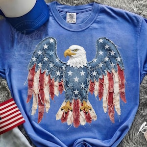 May include: Blue t-shirt featuring a distressed American flag eagle design. The eagle's wings are made of denim with white stars and red and white stripes. The shirt has a crew neck and short sleeves. The tag reads XL - 78-80.