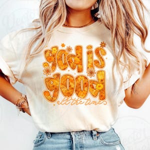 Puede incluir: Camiseta color crema con la frase "God is Good" en un diseño retro floral. El texto es en tonos naranja y amarillo, con "all the time" debajo. La camiseta tiene mangas cortas y cuello redondo.