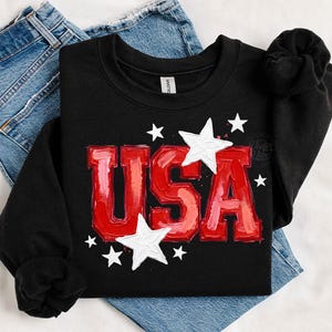 May include: A black long-sleeve crewneck sweatshirt featuring the text USA in a bold, red, textured paint-style font. The design is accented with several white stars of varying sizes scattered around the lettering, creating a patriotic graphic.