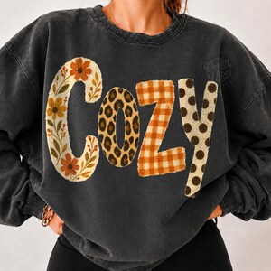 Puede incluir: Una sudadera de cuello redondo gris carb&oacute;n con la palabra COZY impresa en el pecho. Cada letra tiene un patr&oacute;n distinto: la C es floral, la O es de estampado de leopardo, la Z es de cuadros naranjas y la Y tiene lunares marrones sobre crema.