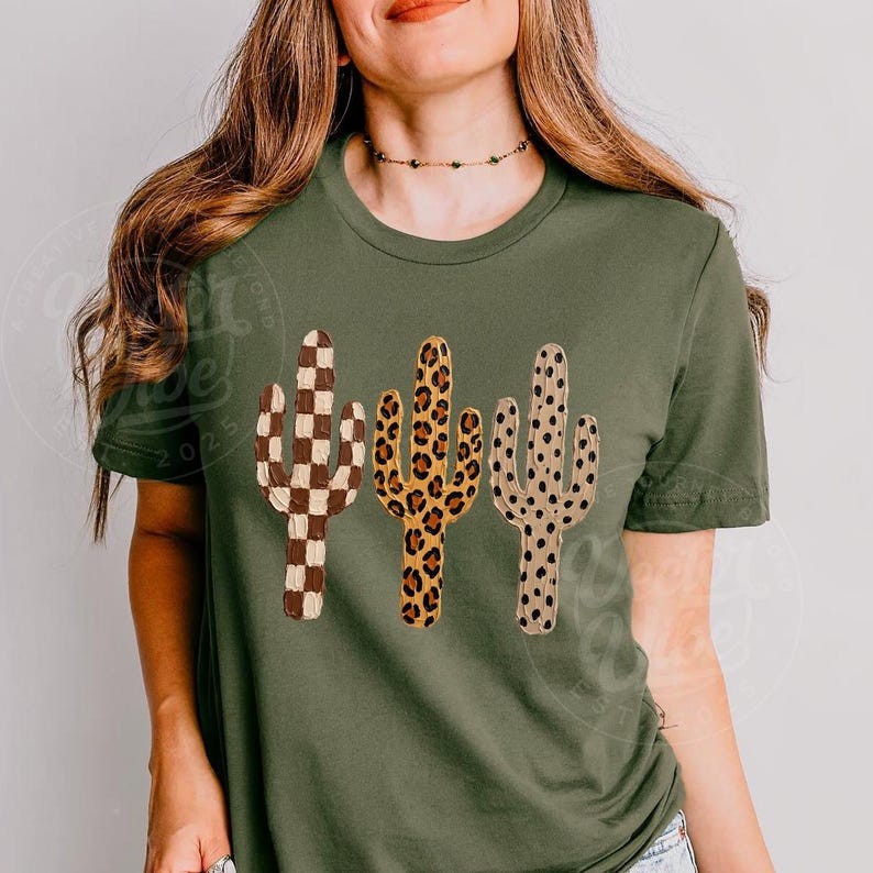 May include: An olive green t-shirt with three cactus designs. The cacti feature a checkered pattern, a leopard print, and a dotted pattern. This casual t-shirt is suitable for everyday wear and is a simple, stylish garment.