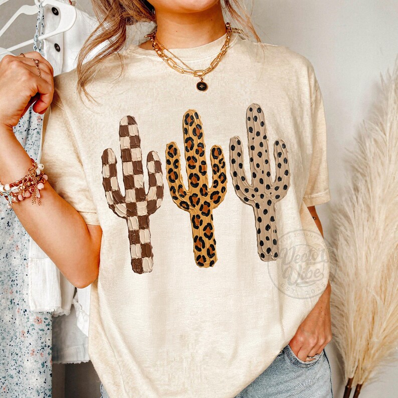 May include: A cream-coloured t-shirt with three cactus designs. The cacti feature a brown and white checkerboard, a leopard print, and a dotted pattern. The shirt is a casual style, suitable for everyday wear.