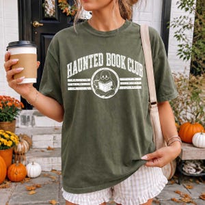 Puede incluir: Una camiseta de manga corta color verde oliva con un estampado gr&aacute;fico blanco que dice "HAUNTED BOOK CLUB" sobre un emblema circular de un fantasma con gafas leyendo un libro y sosteniendo una taza, combinada con pantalones cortos claros con volantes.