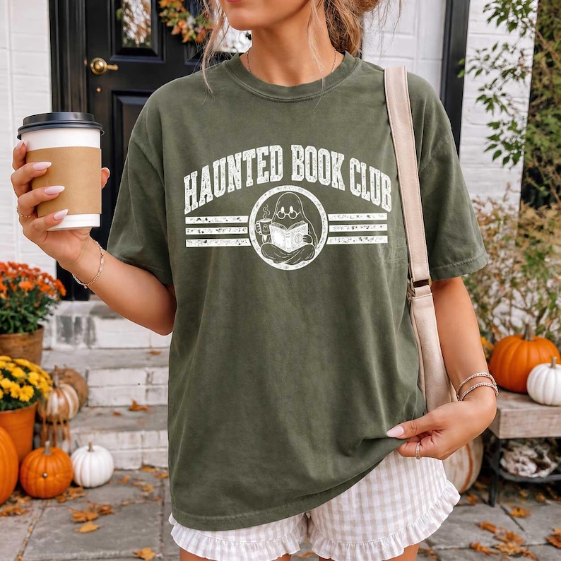 Puede incluir: Una camiseta de manga corta color verde oliva con un estampado gr&aacute;fico blanco que dice "HAUNTED BOOK CLUB" sobre un emblema circular de un fantasma con gafas leyendo un libro y sosteniendo una taza, combinada con pantalones cortos claros con volantes.