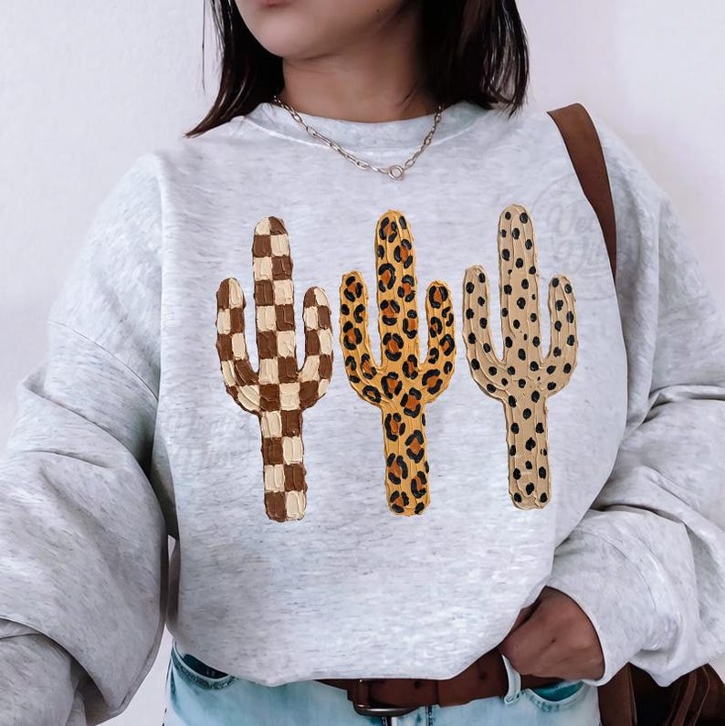 May include: A light grey sweatshirt with three embroidered cactus designs. The cacti feature a brown and white checkerboard, a leopard print, and a tan with brown polka dots. The sweatshirt has a crew neck and long sleeves.