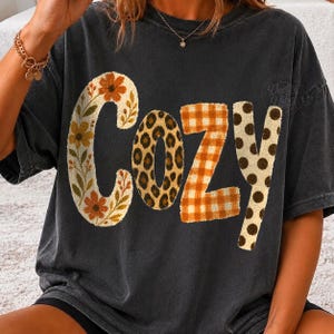 Puede incluir: Una camiseta gris carb&oacute;n de manga corta con la palabra COZY impresa en letras grandes. Cada letra tiene un patr&oacute;n distinto: la C es floral, la O tiene estampado de leopardo, la Z es de cuadros naranjas y la Y es crema con lunares marrones.