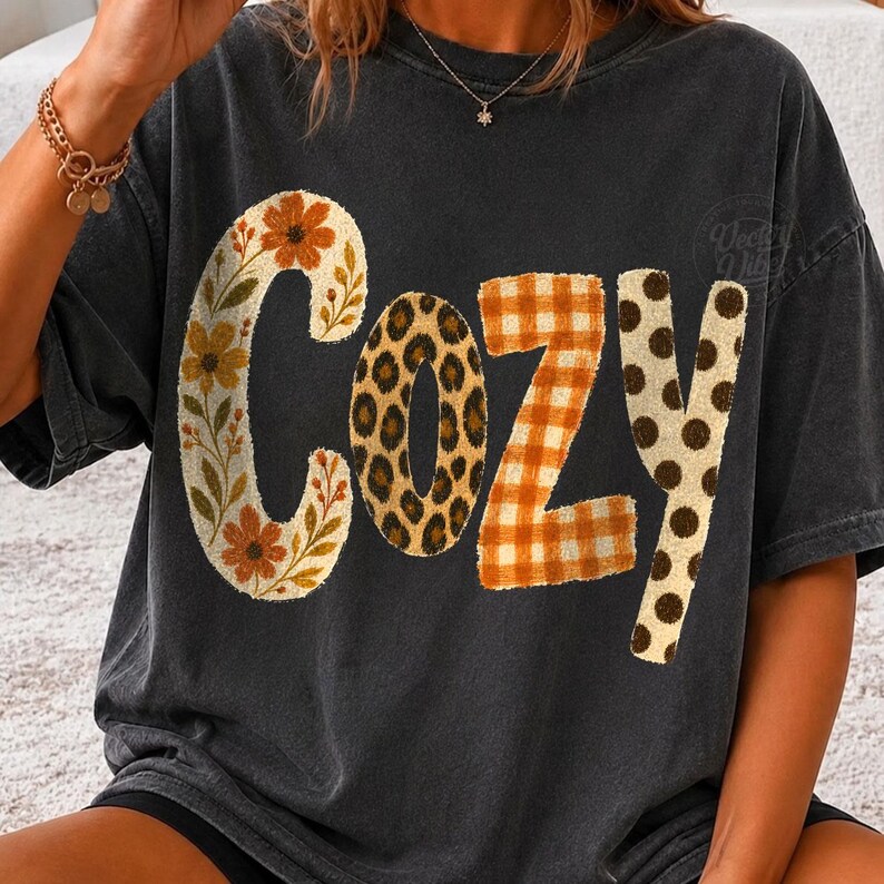 Puede incluir: Una camiseta gris carb&oacute;n de manga corta con la palabra COZY impresa en letras grandes. Cada letra tiene un patr&oacute;n distinto: la C es floral, la O tiene estampado de leopardo, la Z es de cuadros naranjas y la Y es crema con lunares marrones.