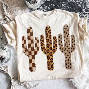 May include: Cream-colored t-shirt featuring three cactus designs. The cacti have unique patterns: a brown and tan checkerboard, a leopard print, and a dotted pattern. The shirt has the text "Vector Vibe" on the left side.