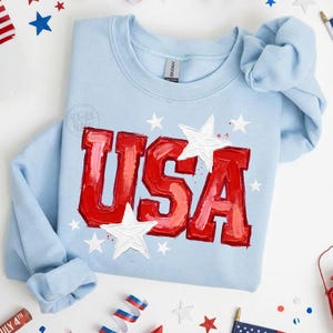 May include: A light blue crewneck sweatshirt featuring the text USA in bold red, textured, paint-like lettering. The design includes several white stars of varying sizes scattered around the letters, creating a patriotic aesthetic on the garment.