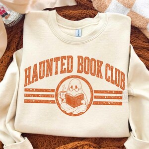 Puede incluir: Una sudadera de cuello redondo color crema con el texto naranja HAUNTED BOOK CLUB en una fuente desgastada. El gr&aacute;fico incluye un emblema circular central de un fantasma con gafas, leyendo un libro abierto y sosteniendo una taza humeante.