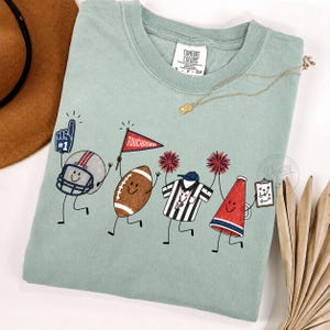 May include: A sage green t-shirt featuring a graphic of four animated football-themed characters: a helmet with a number one foam finger, a brown football holding a touchdown flag, a referee shirt, and a red megaphone holding a play diagram.