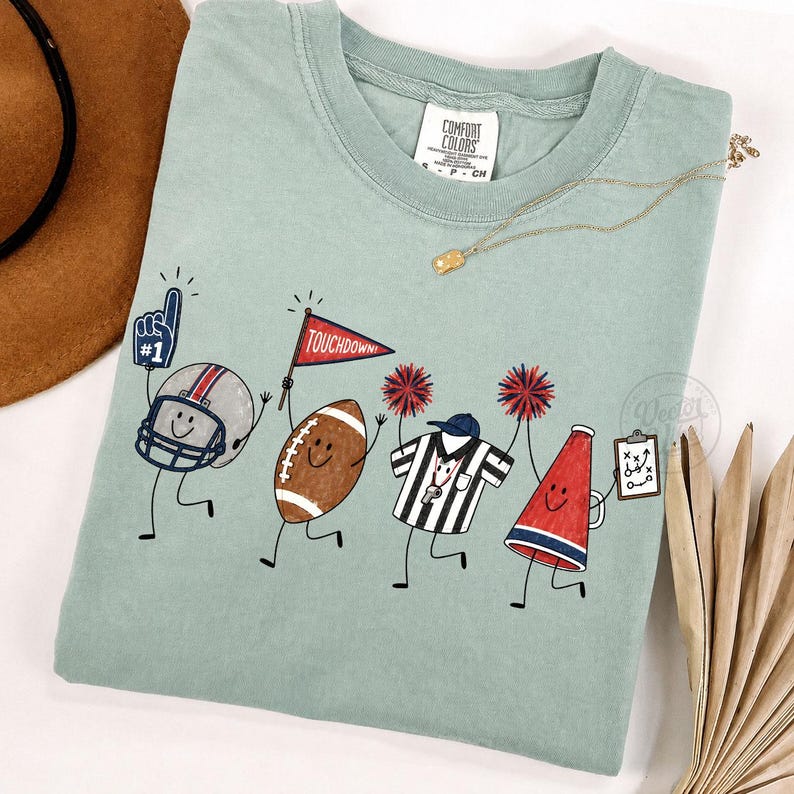 May include: A sage green t-shirt featuring a graphic of four animated football-themed characters: a helmet with a number one foam finger, a brown football holding a touchdown flag, a referee shirt, and a red megaphone holding a play diagram.