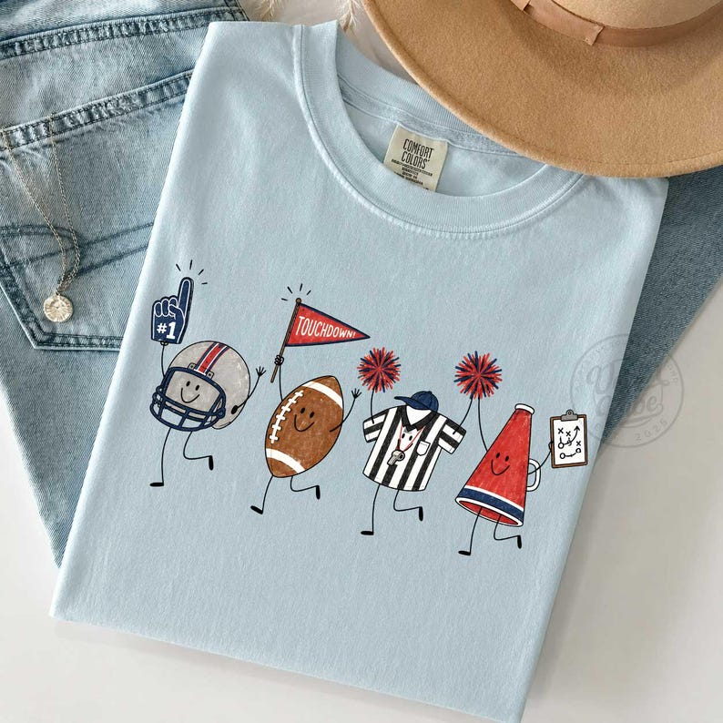 May include: A light blue t-shirt featuring a graphic of four animated football-themed characters: a helmet with a number one foam finger, a football holding a red touchdown flag, a referee in a striped shirt, and a megaphone holding a play sheet.