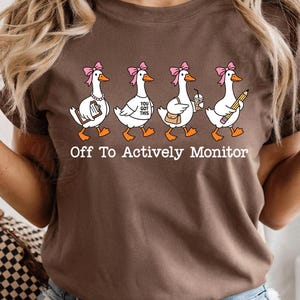 Puede incluir: Una camiseta marrón con un gráfico de cuatro ocas blancas con lazos rosas. Las ocas aparecen con un portapapeles, una camiseta con el texto "YOU GOT THIS", un bolso de hombro con una bebida y un lápiz. El texto "Off To Actively Monitor" aparece impreso debajo.