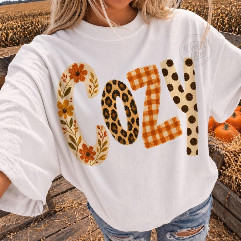 Puede incluir: Una camiseta blanca de cuello redondo con la palabra COZY impresa en letras grandes. La letra C tiene un estampado floral, la O un estampado de leopardo, la Z un cuadro vichy naranja y blanco, y la Y un estampado de lunares marrones.