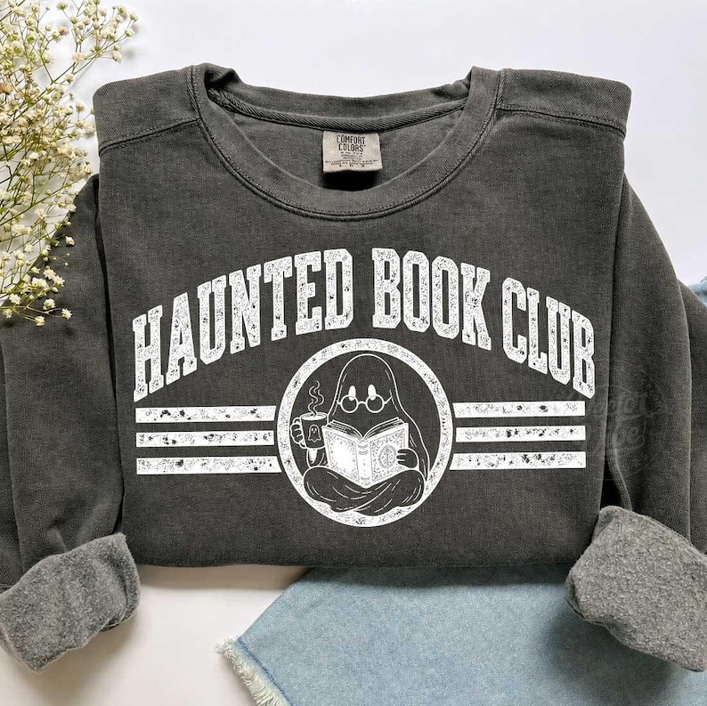 Puede incluir: Una sudadera gris carb&oacute;n de cuello redondo con el texto blanco HAUNTED BOOK CLUB en fuente arqueada. Debajo del texto hay un gr&aacute;fico circular de un fantasma con gafas, sosteniendo una taza humeante y leyendo un libro abierto, flanqueado por l&iacute;neas.