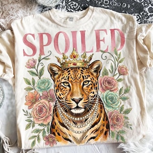 May include: Cream-colored t-shirt with the word "SPOILED" in pink script. The shirt features a leopard wearing a gold crown and pearl necklace, surrounded by pink, peach, and teal roses.
