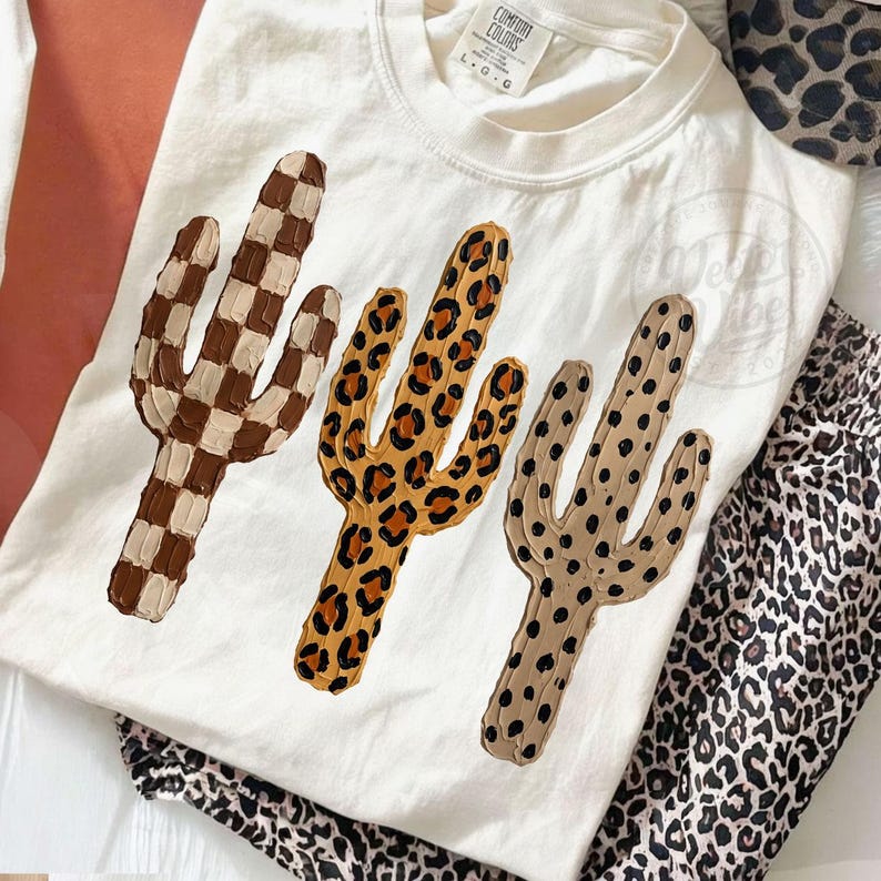 May include: Cream-coloured t-shirt with three cactus designs in brown checkered, leopard print, and dotted tan patterns. The shirt is part of a set with leopard print trousers.