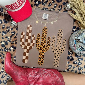 May include: A taupe t-shirt with three cactus designs: a checkered one, a leopard print, and one with black dots. A red baseball cap with "TIMELESS" is visible. The image also shows a red boot and a necklace.