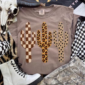 May include: A taupe t-shirt with three cactus designs. The cacti feature a brown and white checkerboard, a leopard print, and a dotted pattern. The shirt is part of a clothing collection.