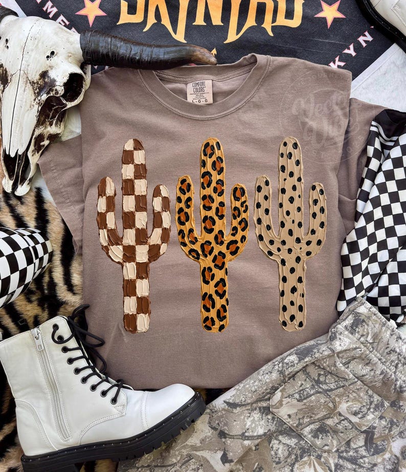 May include: A taupe t-shirt with three cactus designs. The cacti feature a brown and white checkerboard, a leopard print, and a dotted pattern. The shirt is part of a clothing collection.