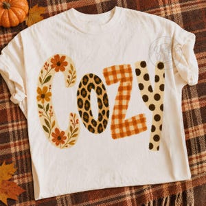 Puede incluir: Una camiseta color hueso con la palabra COZY impresa en letras grandes. La letra C tiene un estampado floral, la O un estampado de leopardo, la Z un patr&oacute;n de cuadros vichy naranja y blanco, y la Y un patr&oacute;n de lunares marrones.