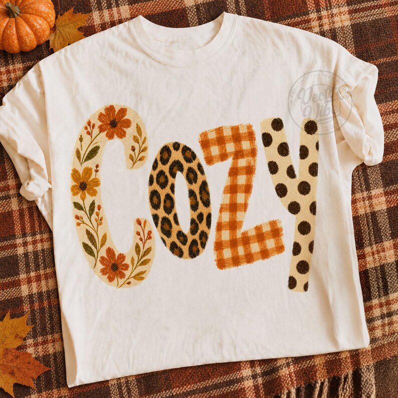 Puede incluir: Una camiseta color hueso con la palabra COZY impresa en letras grandes. La letra C tiene un estampado floral, la O un estampado de leopardo, la Z un patr&oacute;n de cuadros vichy naranja y blanco, y la Y un patr&oacute;n de lunares marrones.