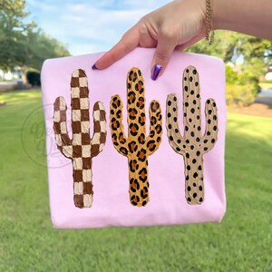 May include: A light pink sweatshirt with three cactus designs. The cacti are in brown and white checkered, leopard print, and tan with black spots. The sweatshirt is held up by a person's hand.
