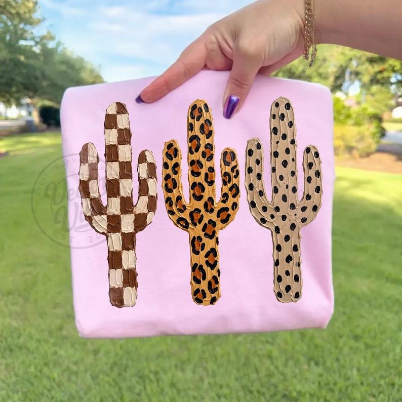 May include: A light pink sweatshirt with three cactus designs. The cacti are in brown and white checkered, leopard print, and tan with black spots. The sweatshirt is held up by a person's hand.
