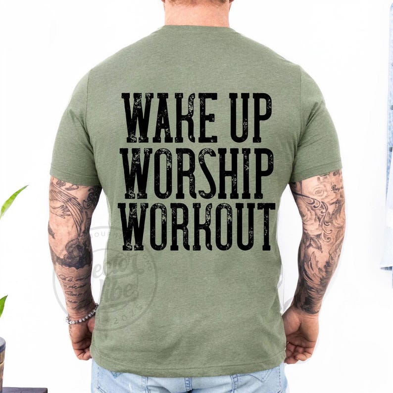 Puede incluir: Camiseta verde oliva con el texto "WAKE UP WORSHIP WORKOUT" en una fuente negra desgastada. La camiseta es de cuello redondo y parece estar hecha de un material suave. La parte trasera de la camiseta es visible.