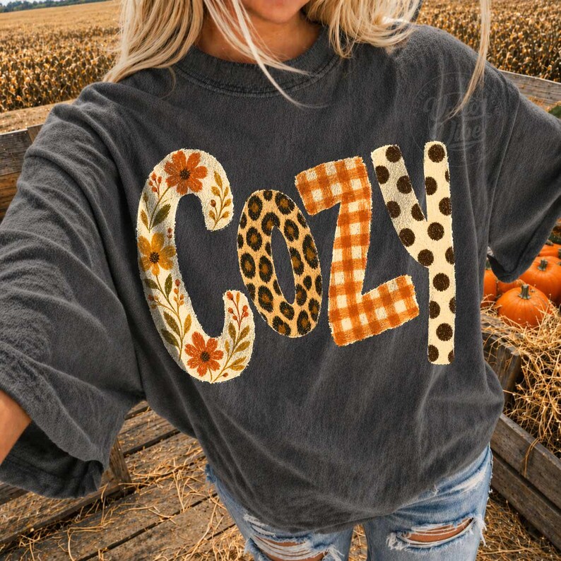 Puede incluir: Una camiseta gris carb&oacute;n de manga corta con la palabra COZY impresa en el frente. Cada letra tiene un patr&oacute;n distinto: la C tiene flores y hojas naranjas, la O un estampado de leopardo, la Z un cuadro naranja y la Y tiene puntos marrones.