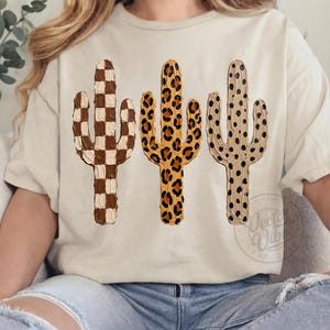 May include: A cream-coloured t-shirt displaying three cactus designs. The cacti feature distinct patterns: a checkerboard, leopard print, and a dotted design. The shirt has a relaxed fit, suitable for casual wear.
