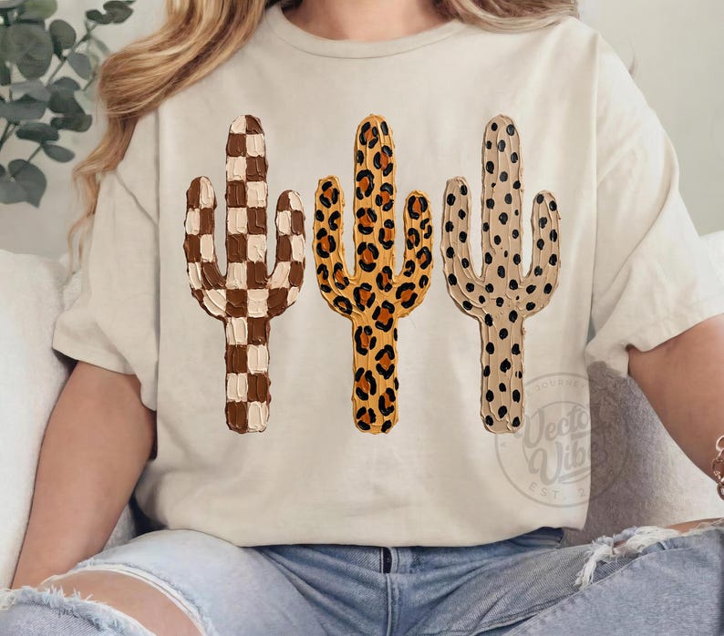May include: A cream-coloured t-shirt displaying three cactus designs. The cacti feature distinct patterns: a checkerboard, leopard print, and a dotted design. The shirt has a relaxed fit, suitable for casual wear.