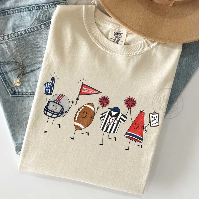 May include: A cream-colored t-shirt featuring a graphic print of four animated football-themed characters: a helmet with a number one foam finger, a football holding a red pennant, a referee shirt, and a megaphone holding a play-calling clipboard.