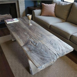 Könnte beinhalten: Rustikaler, rechteckiger Couchtisch aus verwittertem Holz. Der Tisch hat eine natürliche, graue Farbe mit sichtbarer Holzmaserung. Ein Buch und ein Glas stehen auf dem Tisch. Im Hintergrund ist ein beiges Sofa zu sehen.