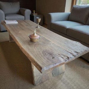 May include: A rustic wooden coffee table with a natural edge, supported by two sturdy wooden legs. The table's surface displays a rich, warm wood grain. A small decorative pot sits on the table, along with a glass and a book. The table is in a living room setting.