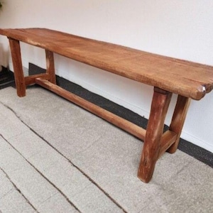 May include: A wooden bench with a natural finish. The bench has a rectangular seat and angled legs. The wood grain is visible, and the bench appears to be handmade. It is a rustic style bench, suitable for indoor or outdoor use.