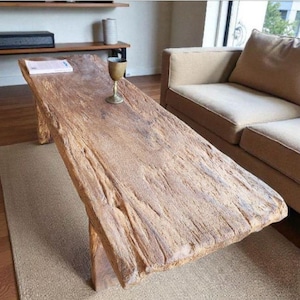 Op de afbeelding: Een rustieke houten salontafel met een natuurlijke, getextureerde afwerking. De tafel heeft een onregelmatige vorm en staat op een neutraal tapijt. Een messing beker staat op de tafel. De tafel is ongeveer 180 cm lang.