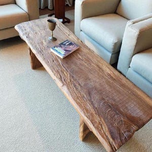 May include: A rustic, rectangular wooden coffee table with a natural edge. A small brass cup and a magazine rest on the table. The table is surrounded by two beige armchairs and a light-colored carpet.