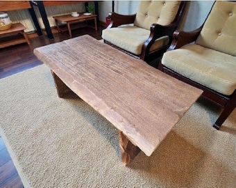Puede incluir: Una mesa de centro rectangular rústica con acabado de madera natural. La mesa tiene una superficie gruesa y texturizada y está sostenida por dos patas anguladas. La mesa está colocada sobre una alfombra beige, con dos sillones al fondo.
