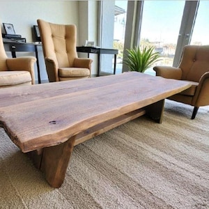 May include: A rustic, rectangular coffee table with a live edge design. The table is made of light brown wood and sits on a sturdy base. Two armchairs with tan upholstery are on either side of the table. The room has a neutral color palette.