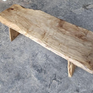 May include: A wooden bench with a live edge. The bench is rectangular with a light brown colour and visible wood grain. It is supported by two triangular legs. The bench is approximately 122 cm long.