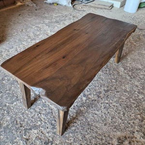 May include: A rectangular coffee table crafted from dark brown wood with a natural live edge along the sides. The table features a smooth, polished surface showing wood grain patterns and is supported by four sturdy, straight wooden legs.